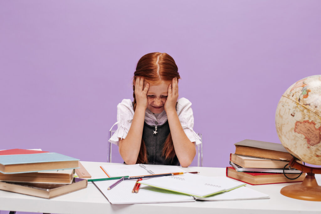 ginger small girl with freckles school uniform holding her head with her hands sitting desk purple background | English Explorers